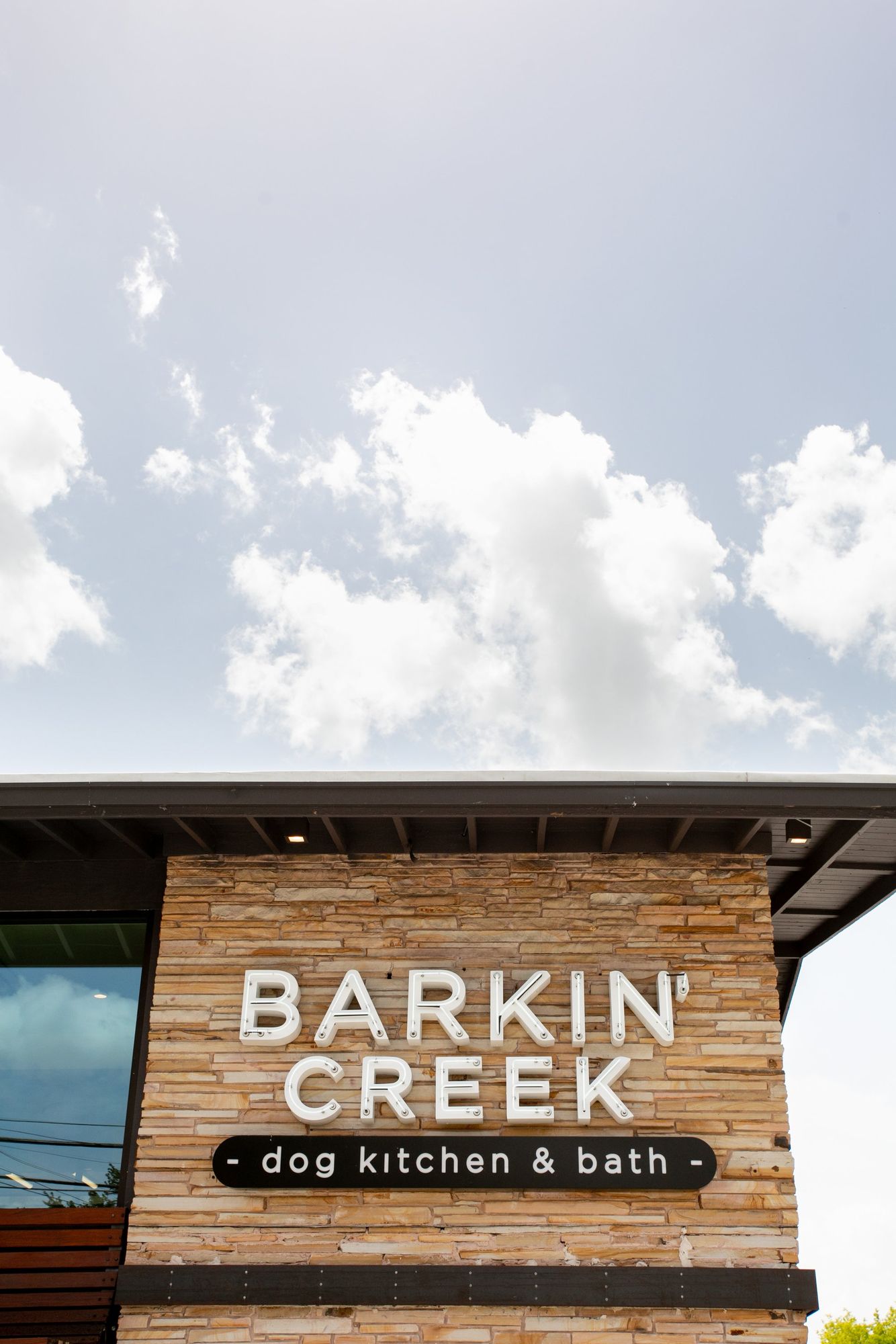 Exterior of Barkin' Creek Dog Kitchen and Bath, a stone-clad single-story commercial building with full storefront glazing and an illuminated dog mascot logo on the right pier