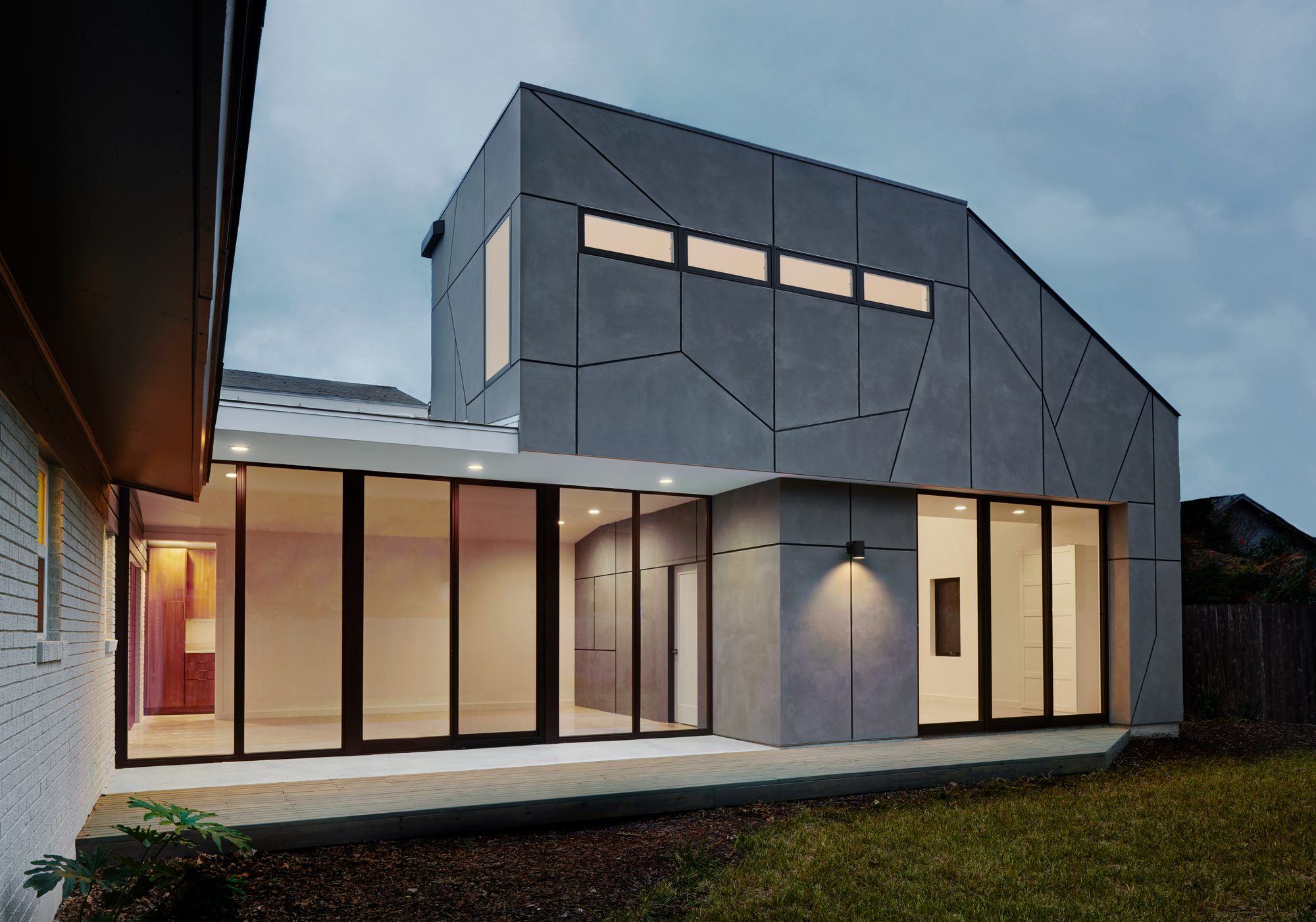 Dusk view of the Choquette House addition from the back yard, showing a faceted grey cementitious-panel volume rising over a glass-walled ground-floor extension, with warm interior light spilling out and the original mid-century ranch visible at left