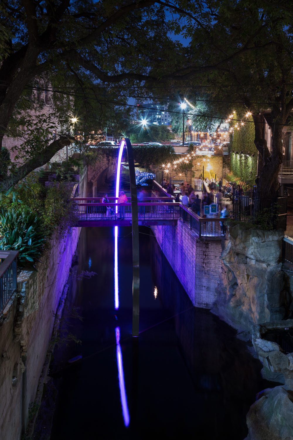 Deep Curiosity installation at night, with a luminous purple arc rising from a bridge over Waller Creek and dipping into the water below, the reflection completing the form as a circle, framed by tree canopy and string lights