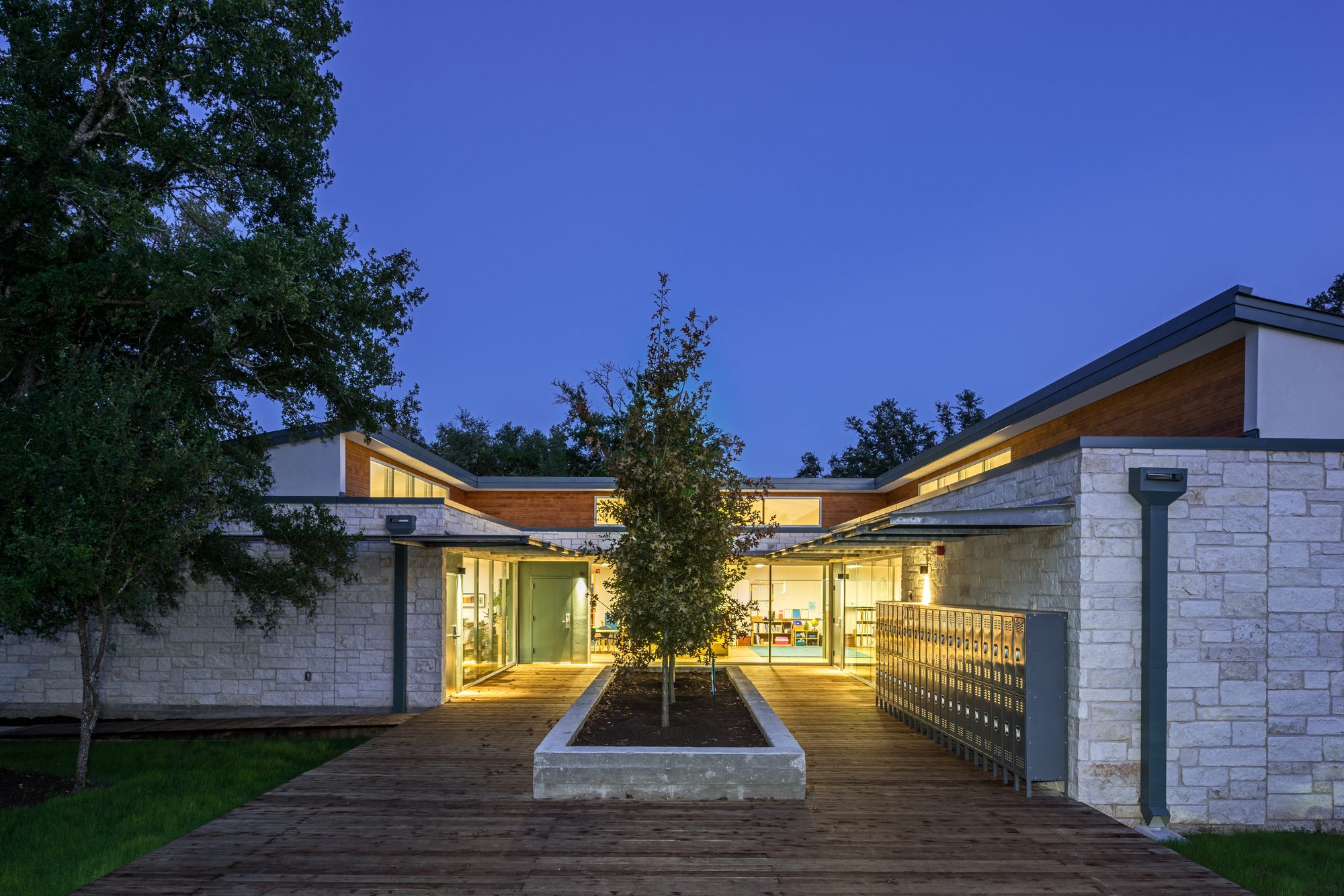 Front exterior of the Headwaters Expansion at twilight, with limestone walls, cedar siding above, a central tree on a wood-deck walkway, and warm interior light spilling out from the entry beyond